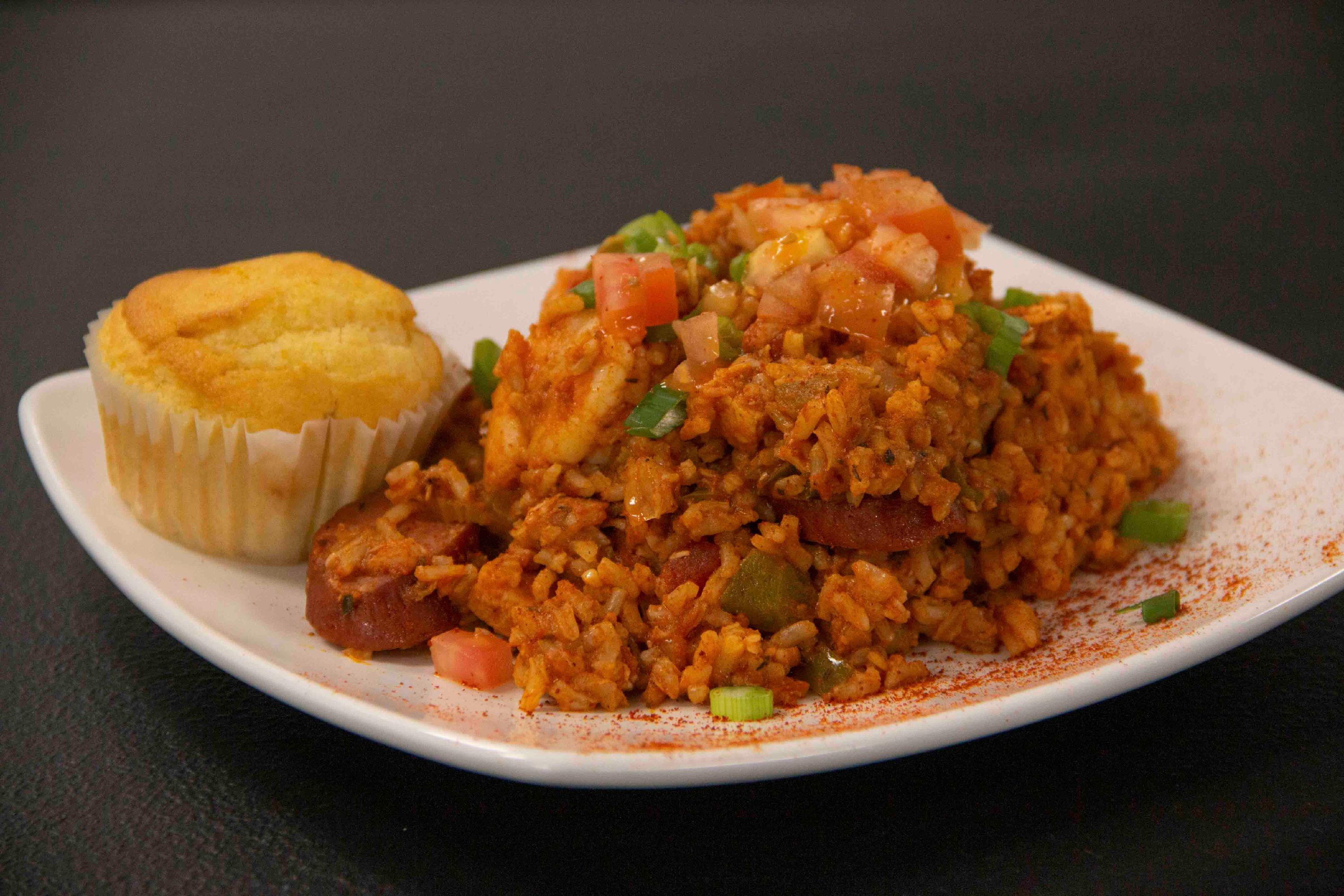 A plate of jambalaya with a small cornbread muffin rests on a dark table